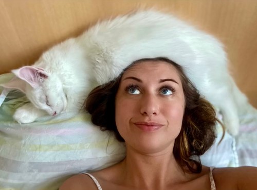 a woman named yuliya minina looking up and smirking at her cat, kefir, who is laying on top of her head as she lays in bed